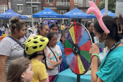 A group of women and young girls look on while a child spins a wheel for a prize at an outdoor event.