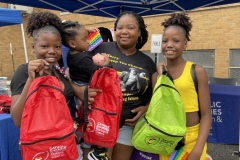 A smiling mother with her three daughters holding up red an green backpacks during an outdoor event.