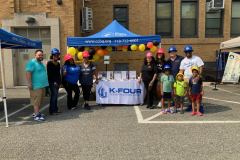 Smiling adults and children in toy construction hats pose at a booth with a K-Four Construction tablecloth at an outdoor event.