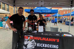 Three smiling people in martial arts uniforms stand at a table with a Tiger Schulman’s tablecloth at an outdoor event.