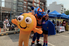 A smiling young woman in a superhero costume poses with a goldfish mascot and an orange and blue dragon mascot in a New York Islanders uniform at an outdoor event.