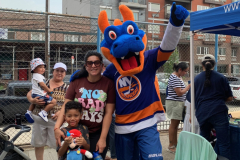 Two mothers and their children pose with a blue and orange dragon mascot in a New York Islanders uniform at an outdoor event.